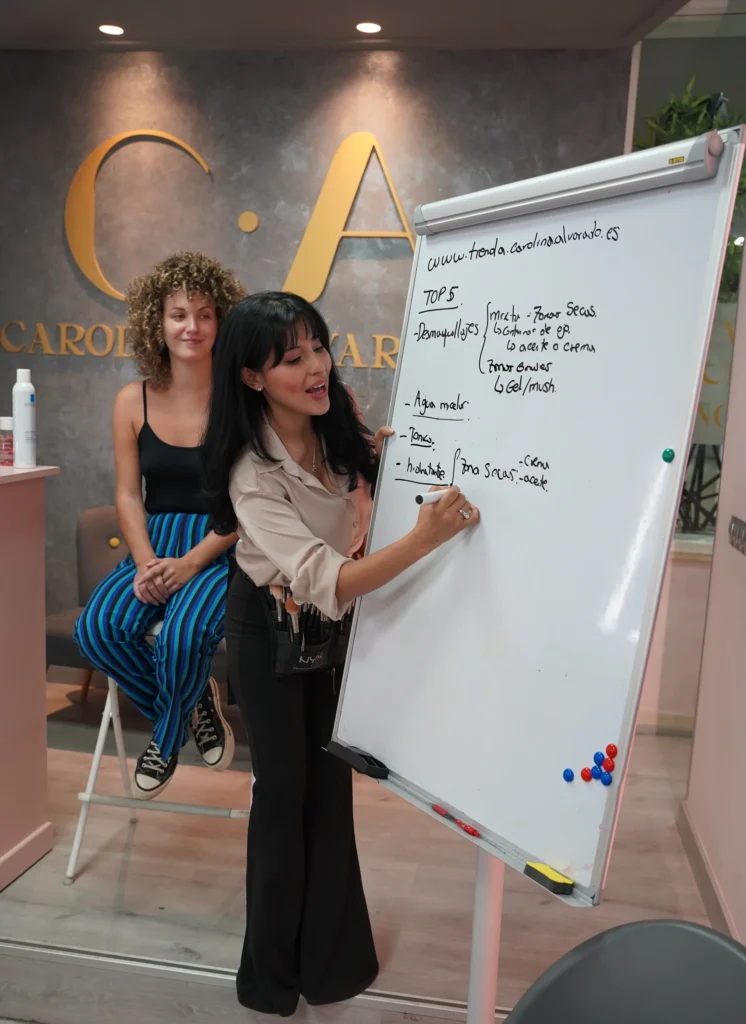 Docente de C•A Academy explicando en una pizarra los pasos de preparación de la piel antes del maquillaje, junto a alumnas atentas a la clase.